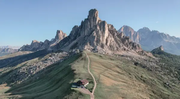 Un refugio alpino solitario en la ladera de una montaña, con un sendero que serpentea hacia él. El paisaje evoca la belleza de **Milano Cortina**, con picos imponentes y cielos despejados.