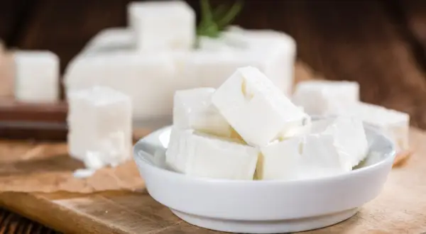 Cubos de queso de leche cruda en un cuenco blanco, con un bloque de queso y una ramita de romero al fondo.