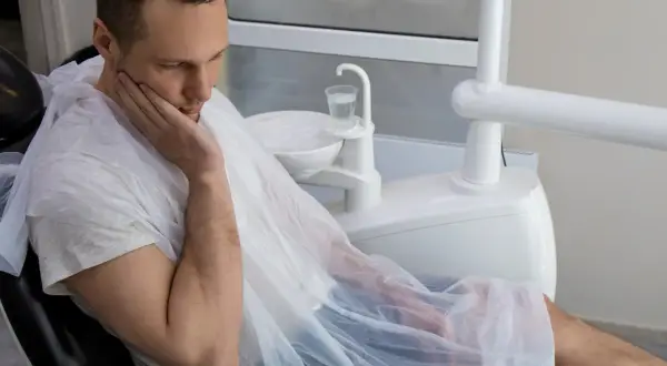 Mężczyzna w fotelu dentystycznym, z ręką na policzku, martwi się o swoje zdrowie. Czy to początek sepsy od zęba?