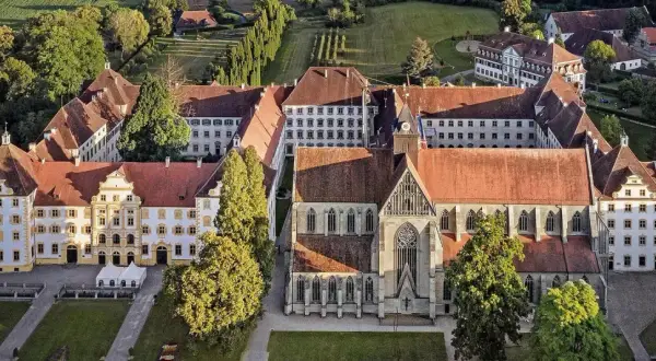 Luftaufnahme eines beeindruckenden schloss im Bodenseekreis mit roten Ziegeldächern, umgeben von grünen Bäumen und gepflegten Gärten.