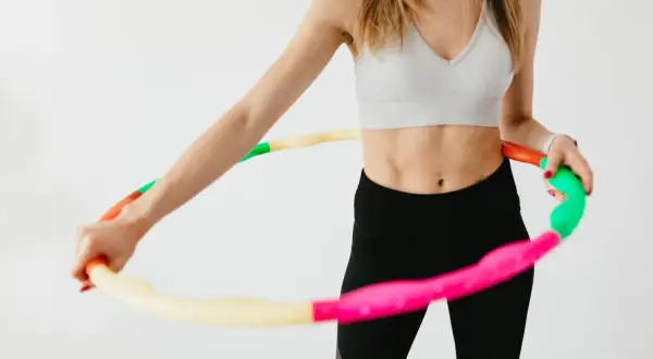 Kobieta w sportowym topie i czarnych legginsach kręci kolorową hula hop. Widać jej wyrzeźbiony brzuch, co sugeruje pozytywne kręcenie hula hop efekty.