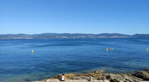Vista desde punta Faxilda. El mar azul se extiende hasta la costa montañosa, con un kayak solitario y boyas amarillas.