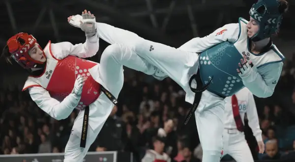 Två taekwondo-utövare i strid. Den ena med röd väst och hjälm, den andra med blå väst och hjälm. Båda utför sparkar.