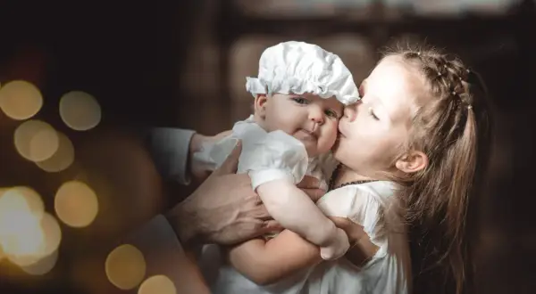 Une sœur embrasse tendrement son bébé, habillé pour baptiser son enfant. Lumières douces et ambiance chaleureuse.