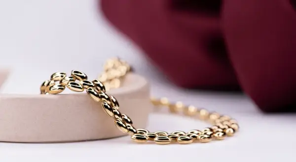 Pulsera de eslabones ovalados en oro, brillando sobre un soporte beige, con un fondo de tela burdeos.