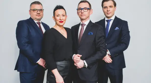 Cztery osoby w garniturach i czarnej bluzce, stojące obok siebie na jasnym tle.