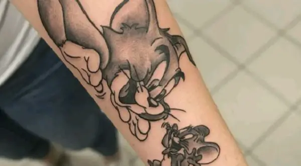 Czarno-biały tatuaż tom i jerry na przedramieniu. Kot Tom próbuje złapać myszkę Jerry, która ucieka.