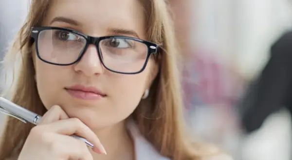 Młoda kobieta w okularach, z długopisem w dłoni, zamyślona. Jej słaby wzrok nie przeszkadza jej w pracy.