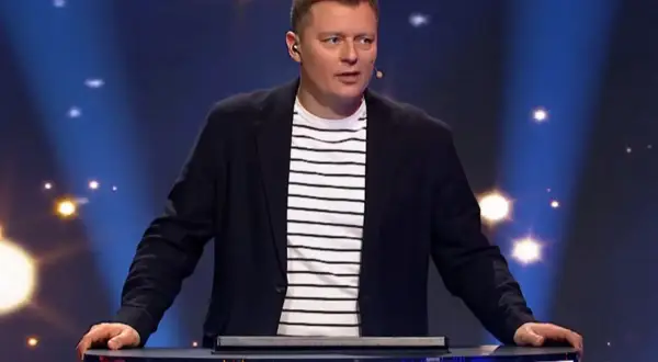 Prowadzący program "Jaka to melodia?" w eleganckim stroju, zastanawia się, ile zarabia zawodowy gracz w Jaka to melodia za jeden odcinek.