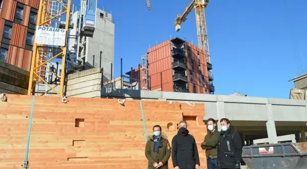 Trois hommes devant un mur en bois, un objet à faire au tour à bois, sur un chantier avec des grues.