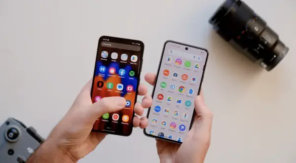 Dwa telefony w dłoniach, jeden z ikoną aparatu, drugi z ikoną aparatu. Który telefon ma lepszy aparat? Obok leży obiektyw aparatu.