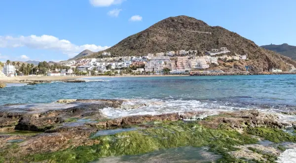 Costa de Cabo de Gata en octubre, con rocas cubiertas de algas verdes y el mar azul. Al fondo, el pueblo blanco se alza en la ladera de una montaña.