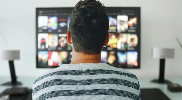 Mężczyzna w pasiastym swetrze ogląda na telewizorze menu aplikacji, gdzie widać okładki filmów i seriali.
