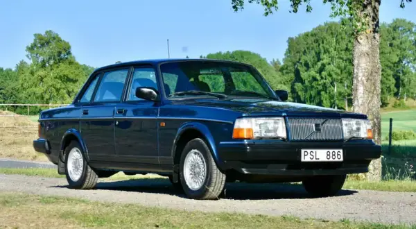 Mörkblå Volvo 240 från 1993 parkerad på en grusväg med grönskande träd och fält i bakgrunden.