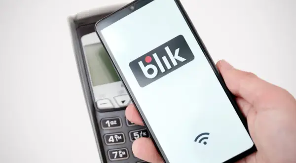 Dłoń trzyma smartfon z logo Blik nad terminalem płatniczym. Szybki przelew na telefon bez obaw o limit.