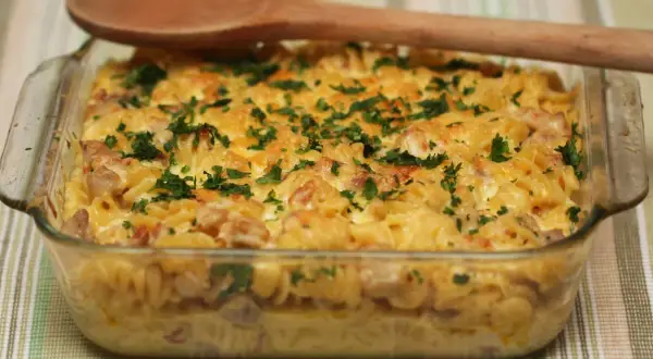 Zapiekanka makaronowa z boczkiem i serem, posypana natką pietruszki, w szklanym naczyniu.