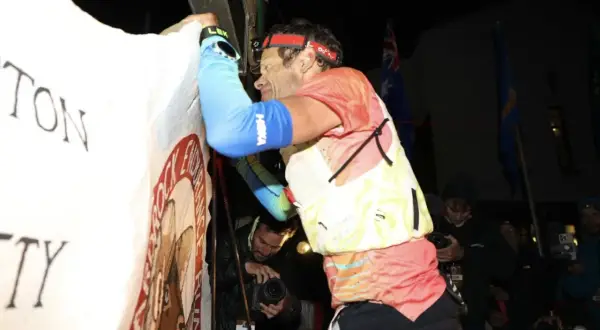 Un coureur, épuisé mais victorieux, est acclamé lors du record Hardrock 100. Des drapeaux flottent derrière lui.