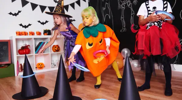 Niñas disfrazadas de bruja y calabaza juegan a lanzar aros en sombreros de bruja, parte de divertidos juegos de Halloween para niños.