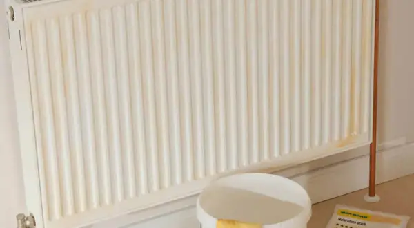 Ein weißer Heizkörper mit gelblichen Flecken, bereit zum heizung streichen. Daneben ein Eimer mit gelben Handschuhen und Malerzubehör.