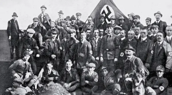 Grupa mężczyzn w mundurach i kapeluszach, z flagą NSDAP w tle. To zdjęcie pokazuje członków partii nazistowskiej, czyli NSDAP.