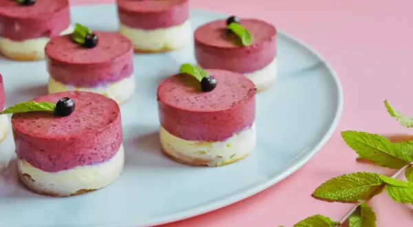 Urocze mini serniczki z jagodami i miętą na jasnoniebieskim talerzu, gotowe do degustacji.
