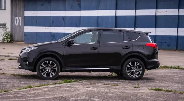 Czarny, używany Toyota RAV4 z salonu, zaparkowany przed budynkiem z niebiesko-białymi pasami.