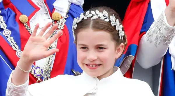 Uśmiechnięta księżniczka Charlotte w tiarze macha ręką, ubrana w białą sukienkę i ozdobne szaty.