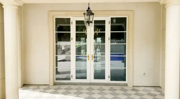 Eleganckie drzwi francuskie z białymi ramami i złotymi klamkami, prowadzące do ogrodu z basenem.