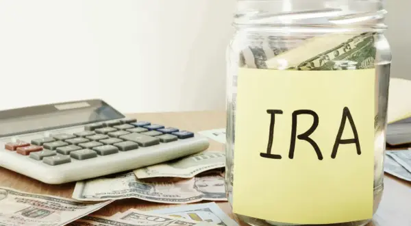 A glass jar labeled "IRA" sits on a table with US dollar bills and a calculator, symbolizing financial planning and the potential for an inherited IRA.