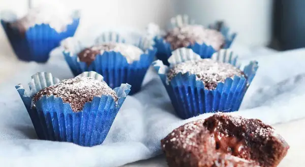 Kladdkakemuffins recept: Mjuka chokladmuffins i blåa pappersformar, pudrade med florsocker. En är uppskuren och visar en härligt kladdig insida.