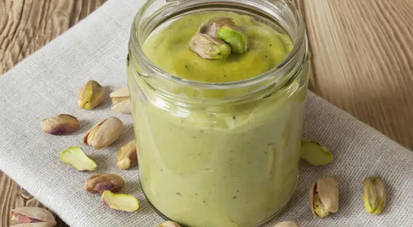 Tarro de crema pistacho casera, decorado con pistachos enteros y trozos, rodeado de más pistachos sobre tela y madera.