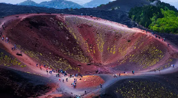 Grupa turystów wędruje po kraterze wulkanu Etna, podziwiając jego czerwoną i czarną, wulkaniczną ziemię porośniętą mchem.