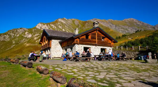 Un rifugio di montagna in pietra e legno, con persone sedute ai tavoli all'aperto, circondato da prati verdi e montagne imponenti sotto un cielo azzurro.