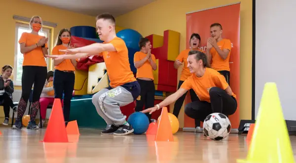 Dzieci i nauczyciele w pomarańczowych koszulkach ćwiczą z piłkami i pachołkami.