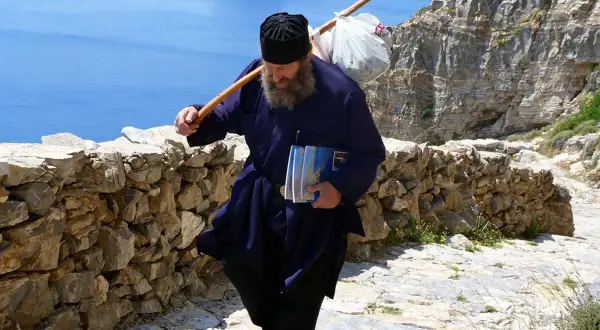 En munk vandrar uppför en stenstig på Amorgos, Grekland, med en stav över axeln och böcker i handen.