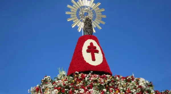 Imagen de una figura religiosa sobre un gran manto de flores rojas y blancas, con un símbolo de cruz. Se celebra una **fiesta** con **pilares** florales.