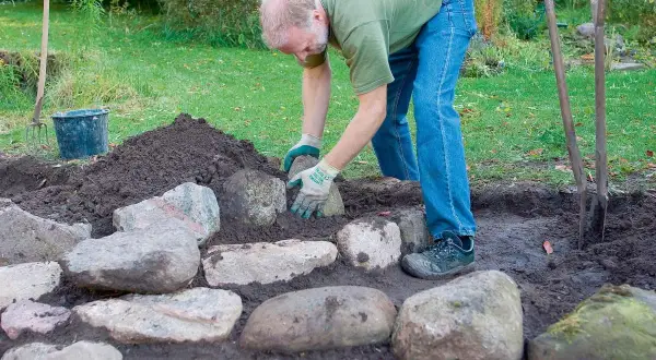 Mężczyzna układa kamienie, tworząc skalniak w ogrodzie. W tle kopiec ziemi i narzędzia ogrodnicze.
