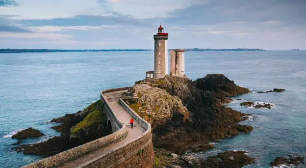 Une personne en manteau rouge marche sur la route des phares, un chemin sinueux menant à un phare sur une île rocheuse.
