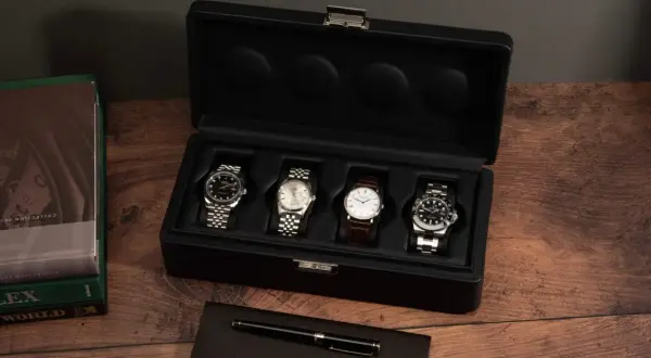 Una caja de reloj negra abierta muestra cuatro relojes de lujo. A la izquierda, libros de Rolex y un bolígrafo.