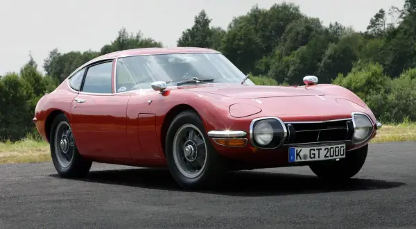 Klasyczny, czerwony Toyota 2000GT, jeden z najbardziej ikonicznych japońskich samochodów sportowych, prezentuje swoje eleganckie linie na tle zieleni.