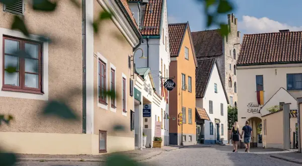 Ett par promenerar hand i hand på en kullerstensgata i Visby, förbi charmiga hus och skyltar som indikerar B&B Gotland och Strand Hotel.