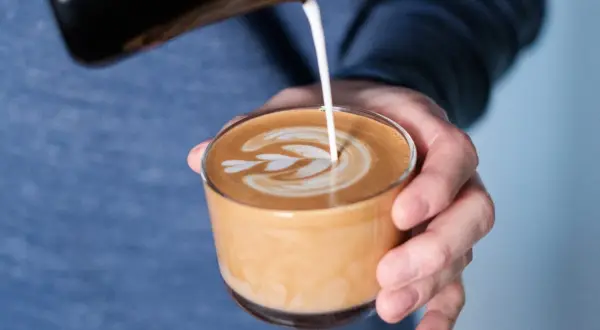 En hand häller mjölk i en kopp för att skapa ett vackert mönster på en flat white.