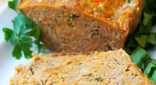 Pyszna pieczeń z mięsa mielonego, pokrojona w plastry, z widocznymi kawałkami marchewki i ziół, udekorowana natką pietruszki.