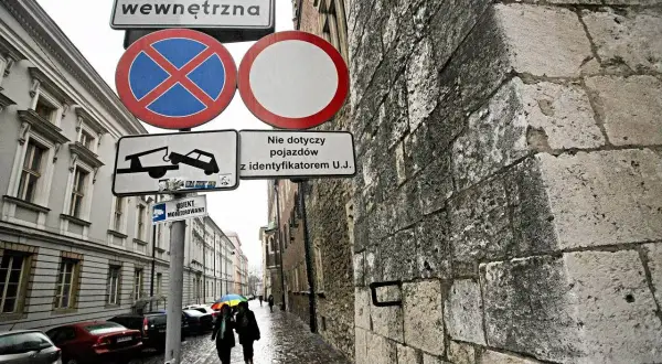 Znaki drogowe informują o zakazie zatrzymywania się i postoju na drodze wewnętrznej. W tle widać ulicę z samochodami i przechodniów.