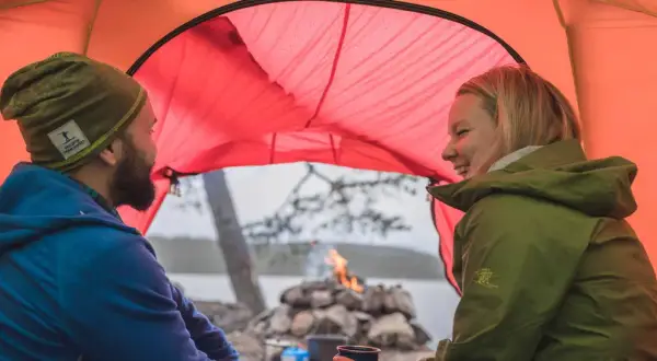 Ett par skrattar inne i ett orange tält. Utanför syns en sjö, träd och en lägereld. Perfekt för att campa i skogen.