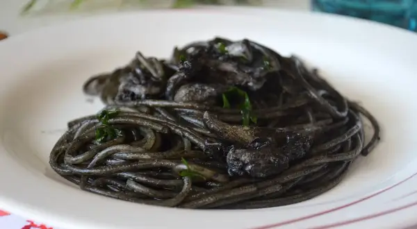 Spaghetti al nero di seppia, un piatto intenso e saporito, servito su un piatto bianco con guarnizioni verdi.