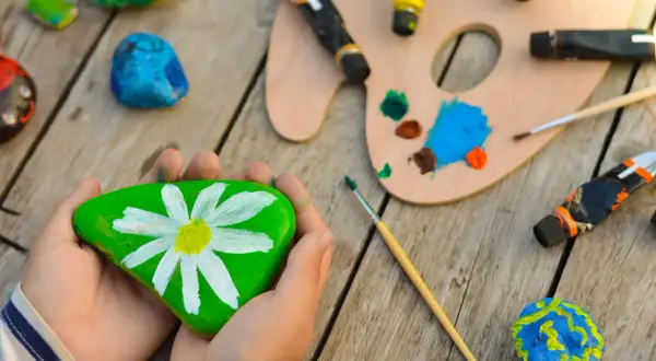 Kreative Ideen zum Steine bemalen für Kinder: Ein grüner Stein mit Gänseblümchen, bunte Farben und Pinsel laden zum Malen ein.