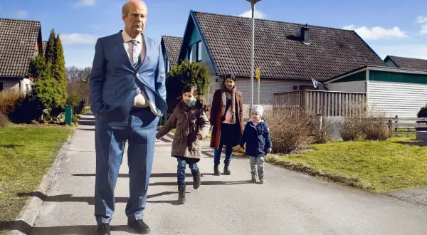 En man i kostym går med en familj i ett villaområde, en scen som kan passa i svenska komedier.