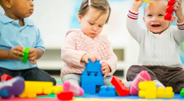 Three toddlers engage in parallel play, each absorbed in their own colorful blocks. One stacks blue blocks, another holds red and yellow toys, and the third plays with green.