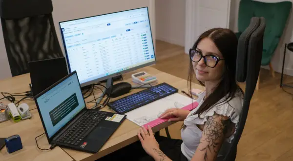 Kobieta w okularach siedzi przy biurku z laptopem i monitorem, na którym widać tabelę danych.